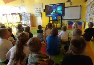 Grupa dzieci siedzi przed ekranem i ogląda spektakl muzyczny.